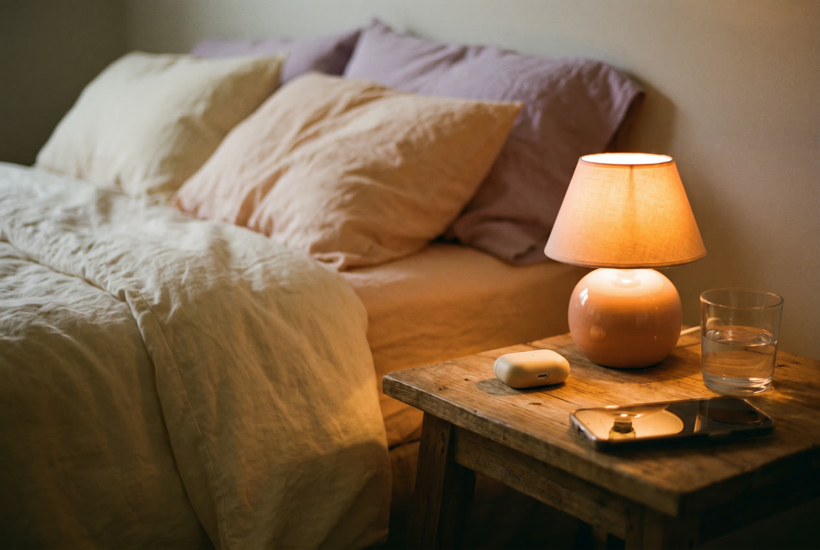 A bedside nightstand at night with a glowing lamp, an earbuds case, a glass of water and a phone face down.